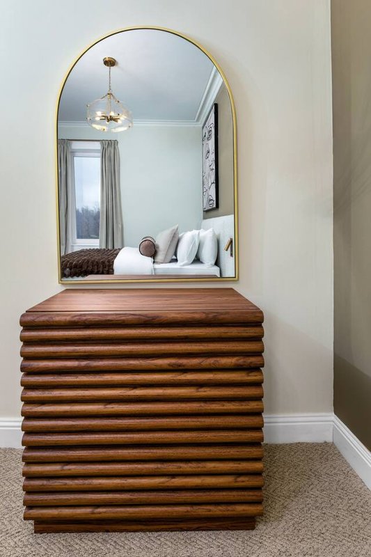 Wooden dresser with arched mirror reflecting bedroom with bed and pendant light