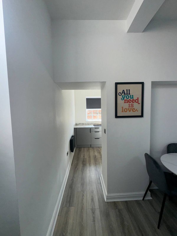Modern hallway with wooden flooring, white walls, framed wall art, and kitchen visible in background