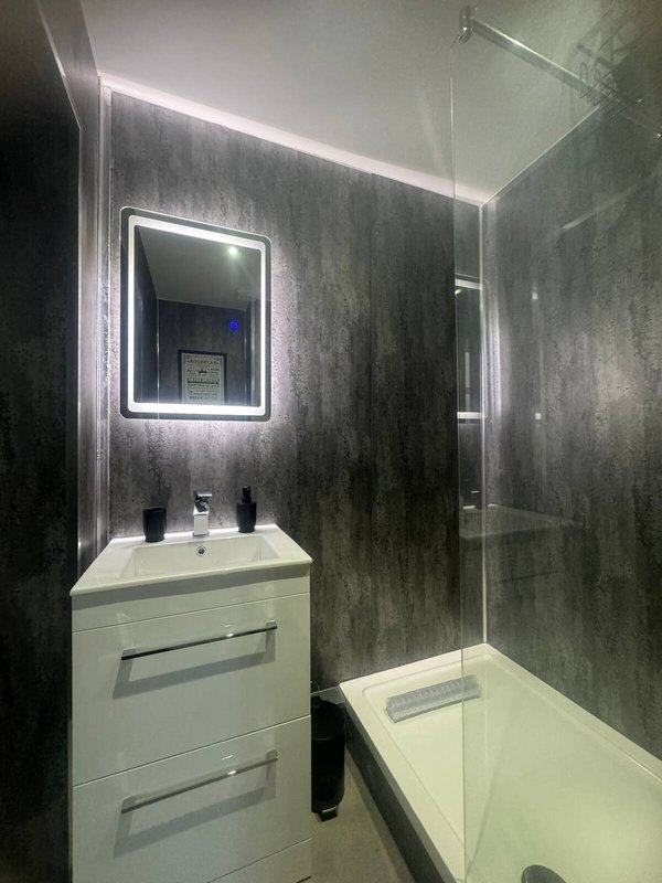 Modern bathroom with illuminated mirror, white vanity unit, dark tile walls and bathtub