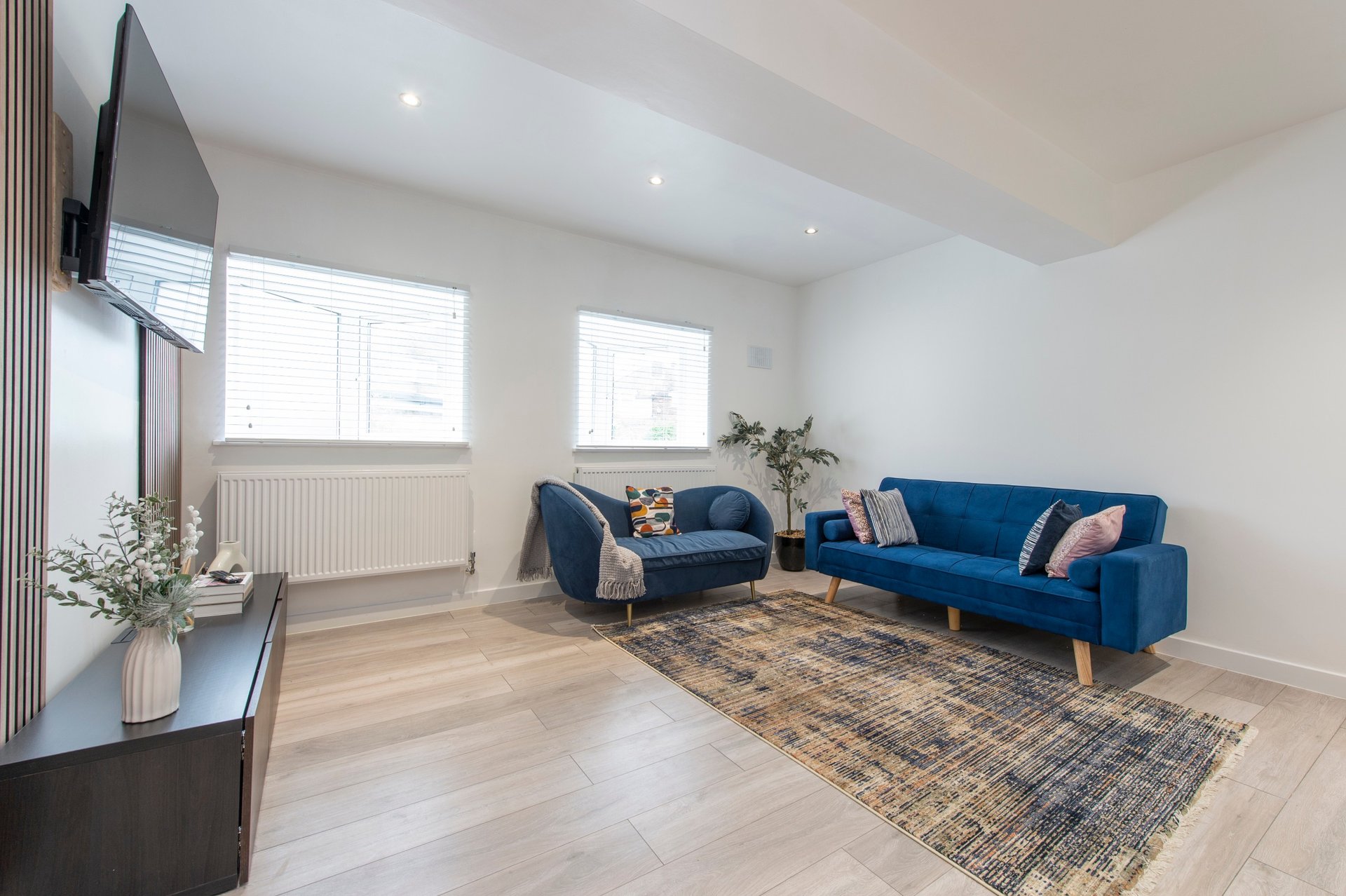 Bright living room with blue sofa and armchair, wooden floor, wall-mounted TV and windows