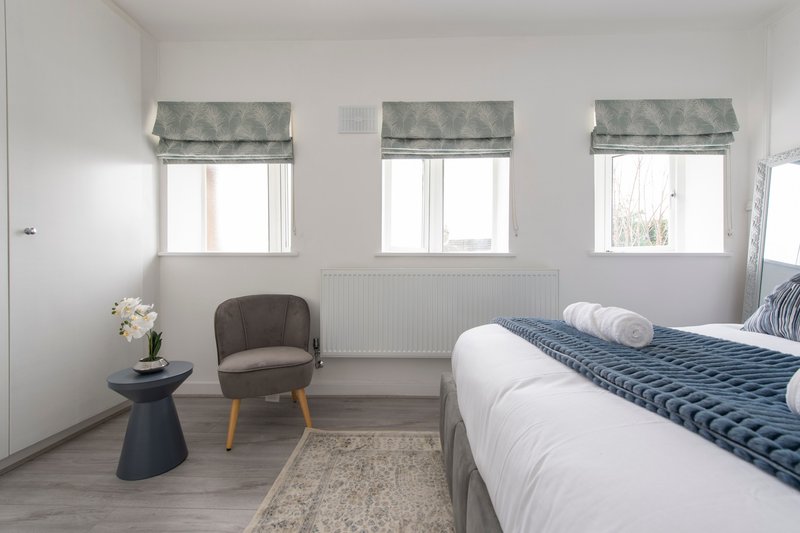 Bright bedroom with double bed, grey armchair, small side table and three windows with roman blinds