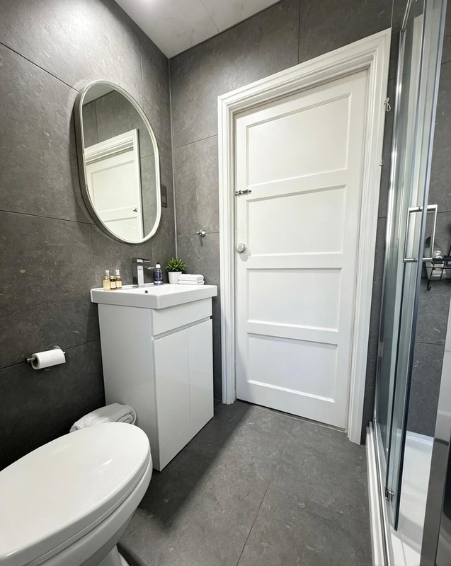 Modern bathroom with grey tile walls, white toilet, sink vanity, oval mirror and shower enclosure