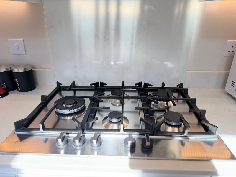 Stainless steel gas cooktop with five burners and glass splashback in kitchen
