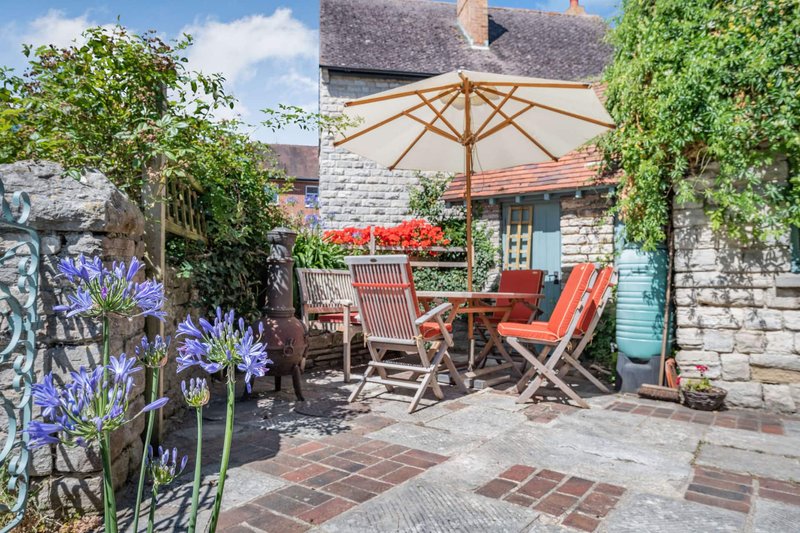Cottage garden patio with seating area, umbrella, flowering plants and ivy-covered walls