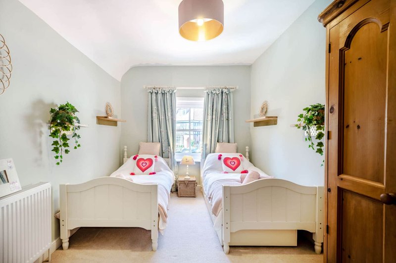 Twin bedroom with two white beds, hanging plants, window with curtains, wooden wardrobe