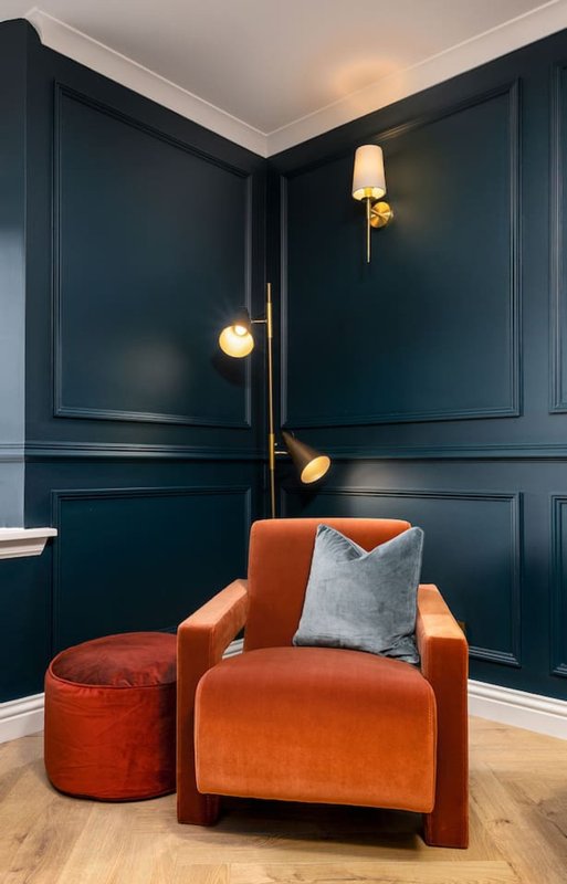 Modern living room with orange armchair, matching ottoman, dark blue panelled walls and brass wall lights