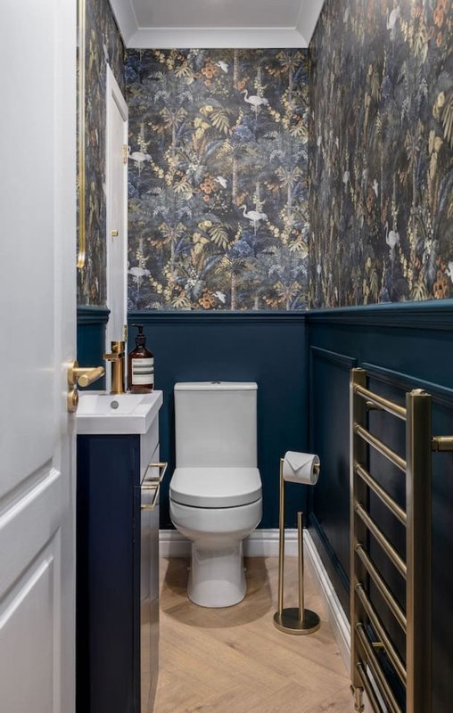 Compact bathroom with white toilet, dark blue paneling, patterned wallpaper and gold fixtures