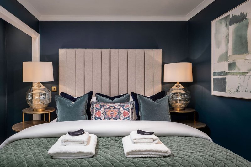 Modern bedroom with upholstered headboard, navy walls, bedside lamps, and neatly folded towels