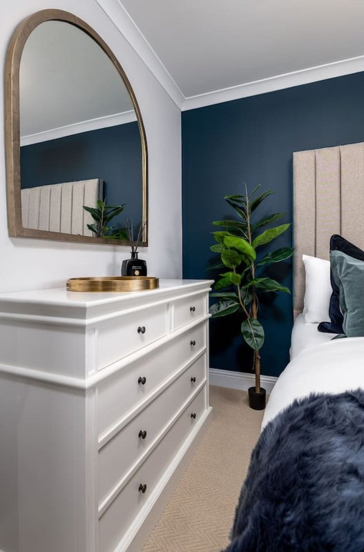 Modern bedroom with white dresser, arched mirror, potted plant and dark blue accent wall