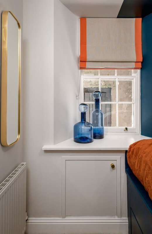 Bright bedroom corner with window, white storage unit, blue glass bottles, and orange roman blind