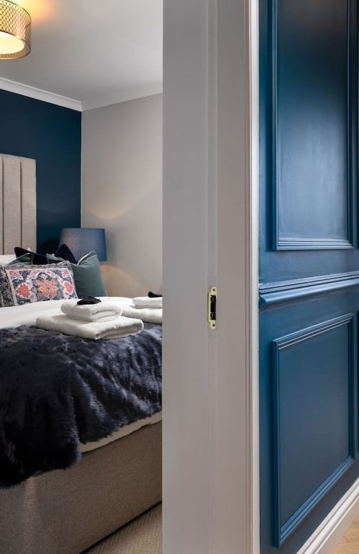 Bedroom with dark blue accent wall, double bed with white pillows and dark bedding, blue door, pendant light