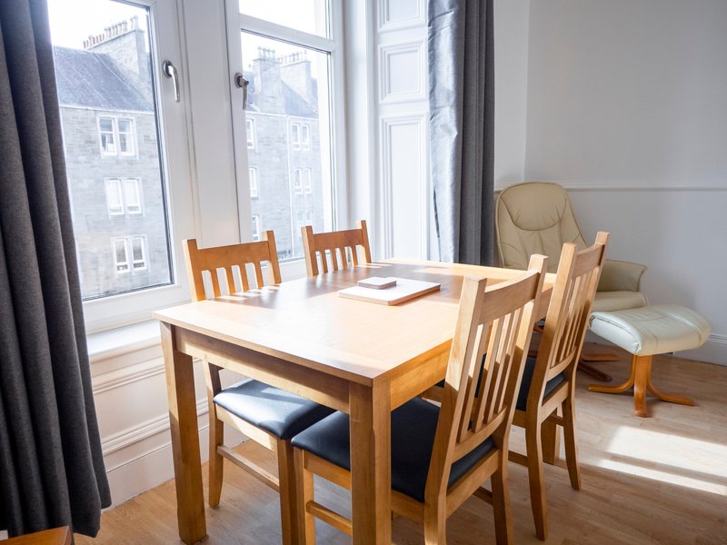 Bright dining area with wooden table, four chairs, large windows and beige armchair
