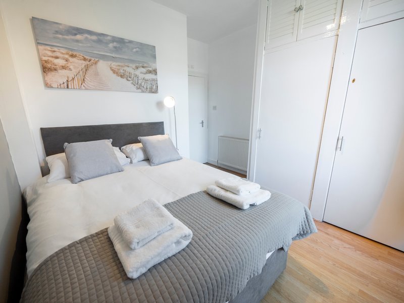 Bright bedroom with double bed, grey headboard, white walls, artwork, and wooden flooring