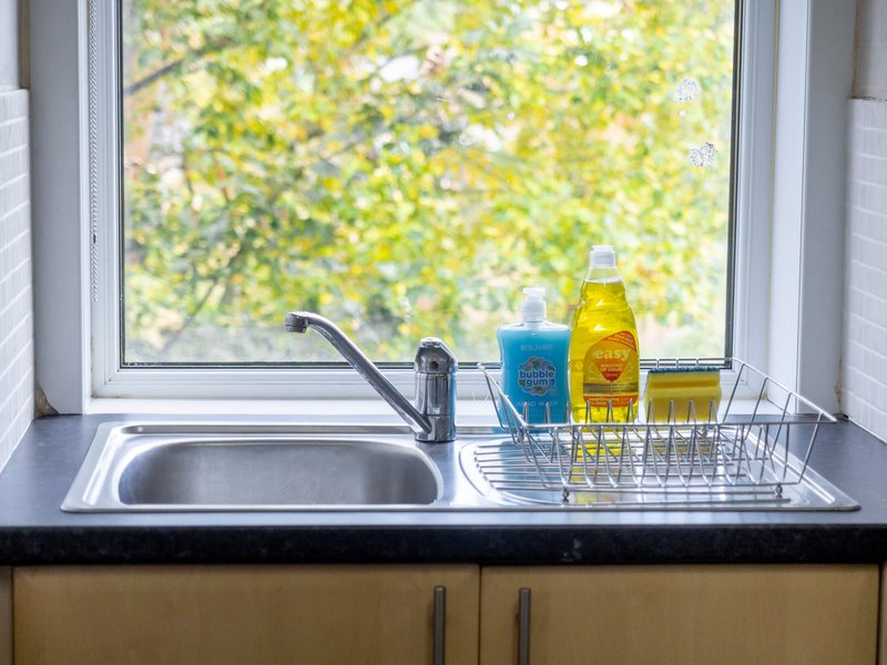 Kitchen sink with tap under window, dish rack with cleaning products and bottles visible