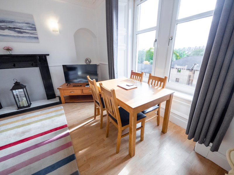 Bright living room with wooden dining table, chairs, fireplace, TV and large windows overlooking street