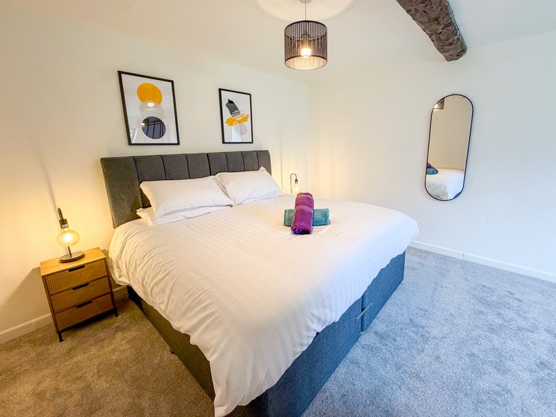 Modern bedroom with grey upholstered bed, white bedding, wooden nightstand, framed wall art, and pendant light