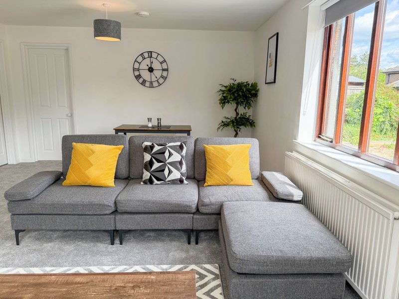 Modern living room with gray L-shaped sofa, yellow cushions, wall clock, and large windows