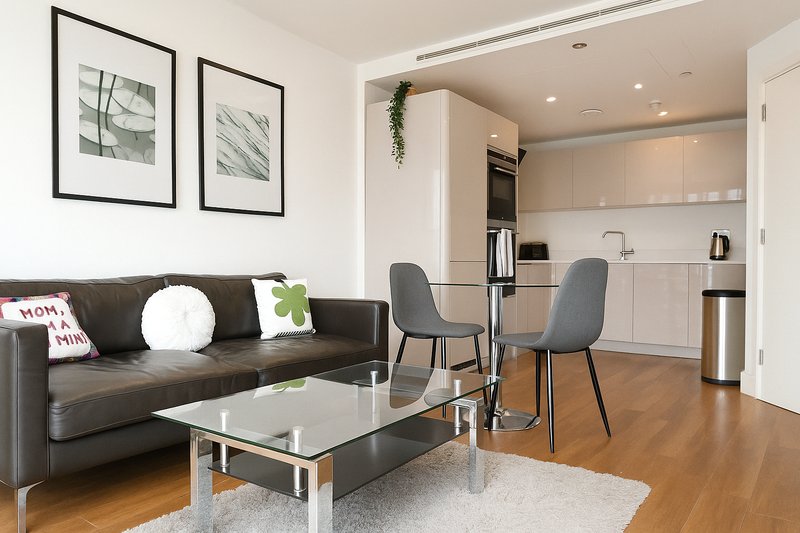 Open-plan living area with dark leather sofa, glass coffee table, dining chairs and modern kitchen visible