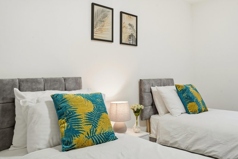 Twin bedroom with gray upholstered headboards, colorful patterned cushions, white bedding, and framed wall art.