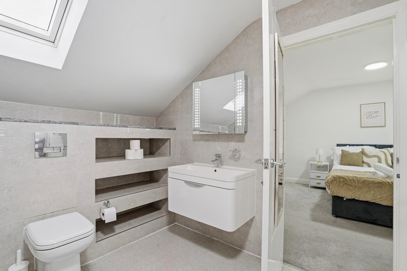 Modern bathroom with white vanity, toilet, mirror, and skylight; bedroom visible through open door