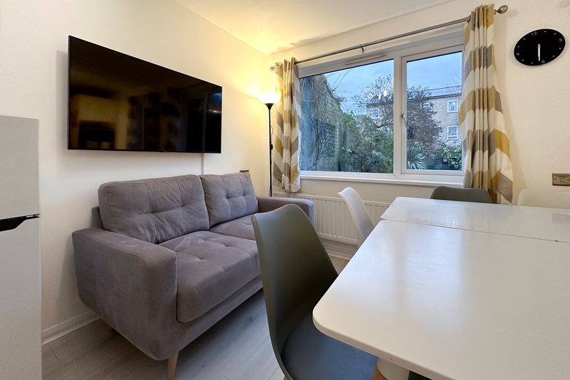Modern living room with gray upholstered sofa, white dining table, wall-mounted TV, and large windows with garden views