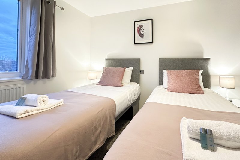 Twin bedroom with two single beds, pink cushions, bedside lamps and window with curtains