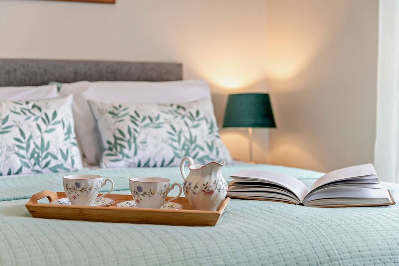 Bedroom with white bedding, patterned cushions, tea service on tray, open book and green table lamp