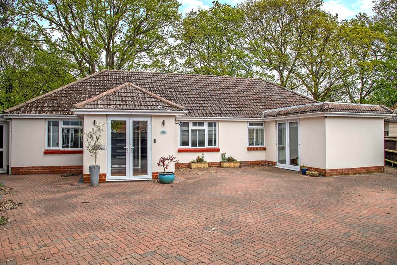 Single-story white bungalow with brick driveway, garden planters, and mature trees
