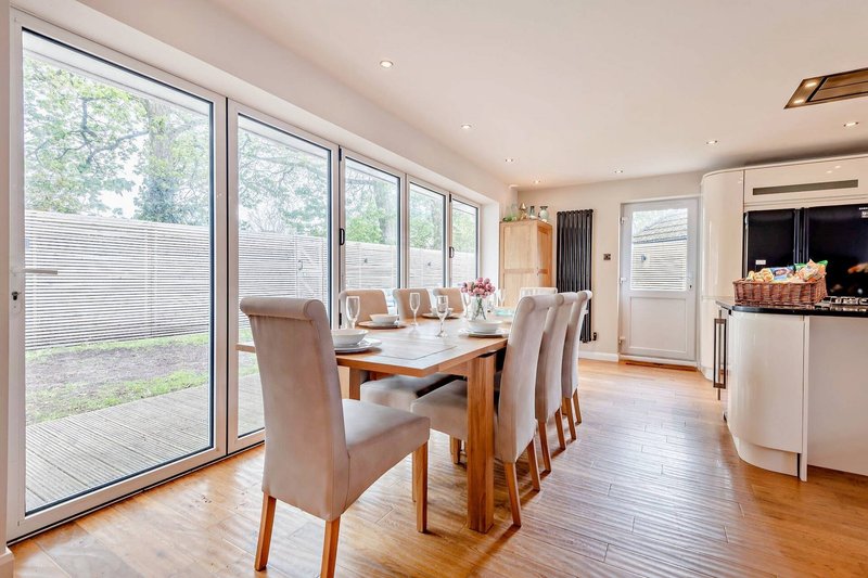 Open-plan dining and kitchen area with wooden table, upholstered chairs, and large glass doors overlooking garden
