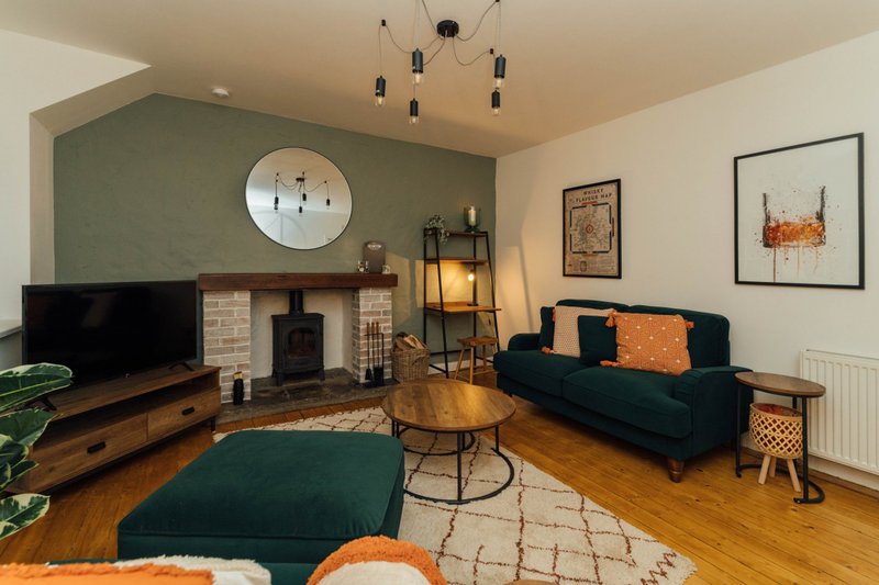 Living room with dark green sofa, wood stove fireplace, wall clock, pendant lighting, and wooden flooring