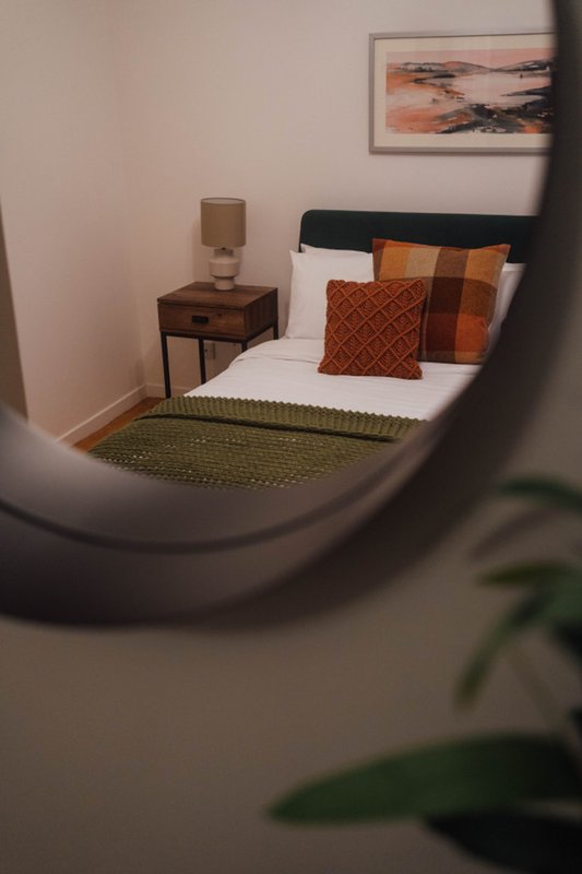 Bedroom with double bed, orange cushions, wooden bedside table, lamp, and framed artwork