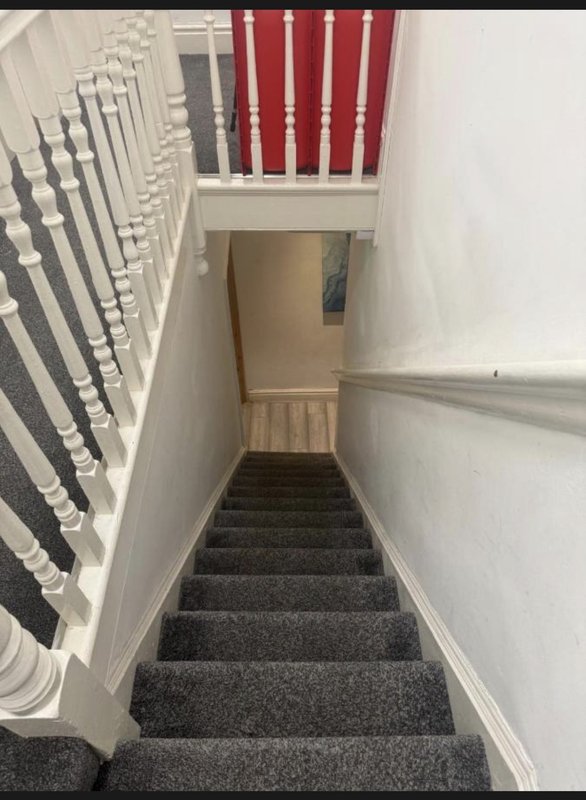 Staircase with gray carpet, white walls, metal handrail, and red curtains visible above