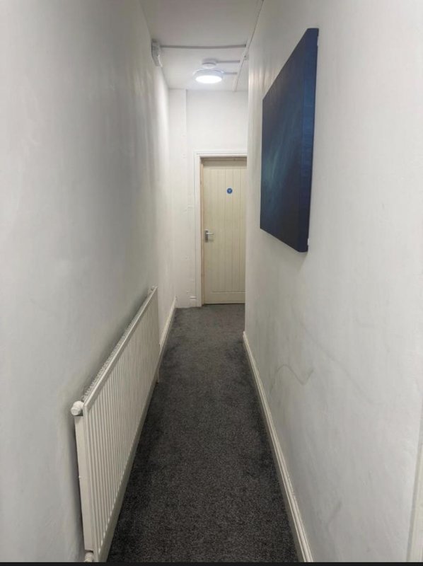 Narrow hallway with white walls, dark carpet, radiator and mounted wall-mounted screen